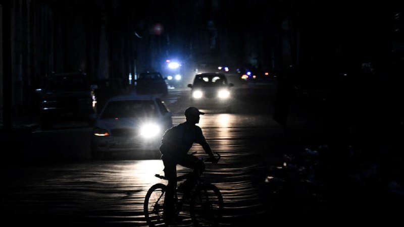 A silhouetted cyclist rides along a dark, wet street at night as several cars approach with headlights illuminating the road.