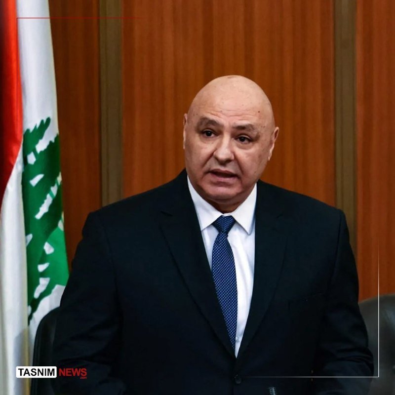 A man in a dark suit and light shirt speaks into a microphone at a podium, with a large banner displaying Arabic script and a portrait of a bearded man behind him.