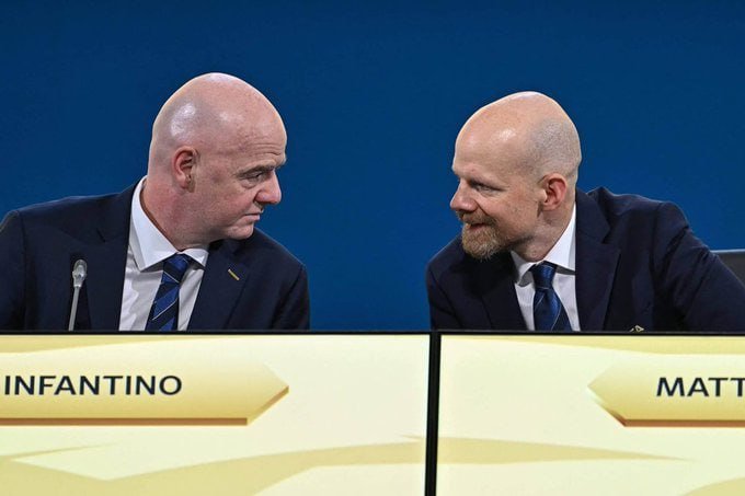Two bald men in dark suits and blue ties sit at a conference table looking at each other, with nameplates reading "INFANTINO" and "MATT" in front of them.