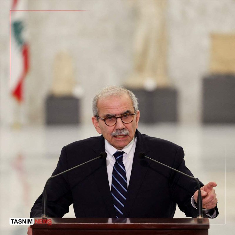 An older man with a mustache and glasses speaks at a podium with two microphones, gesturing with one hand, with a blurred flag visible in the background.