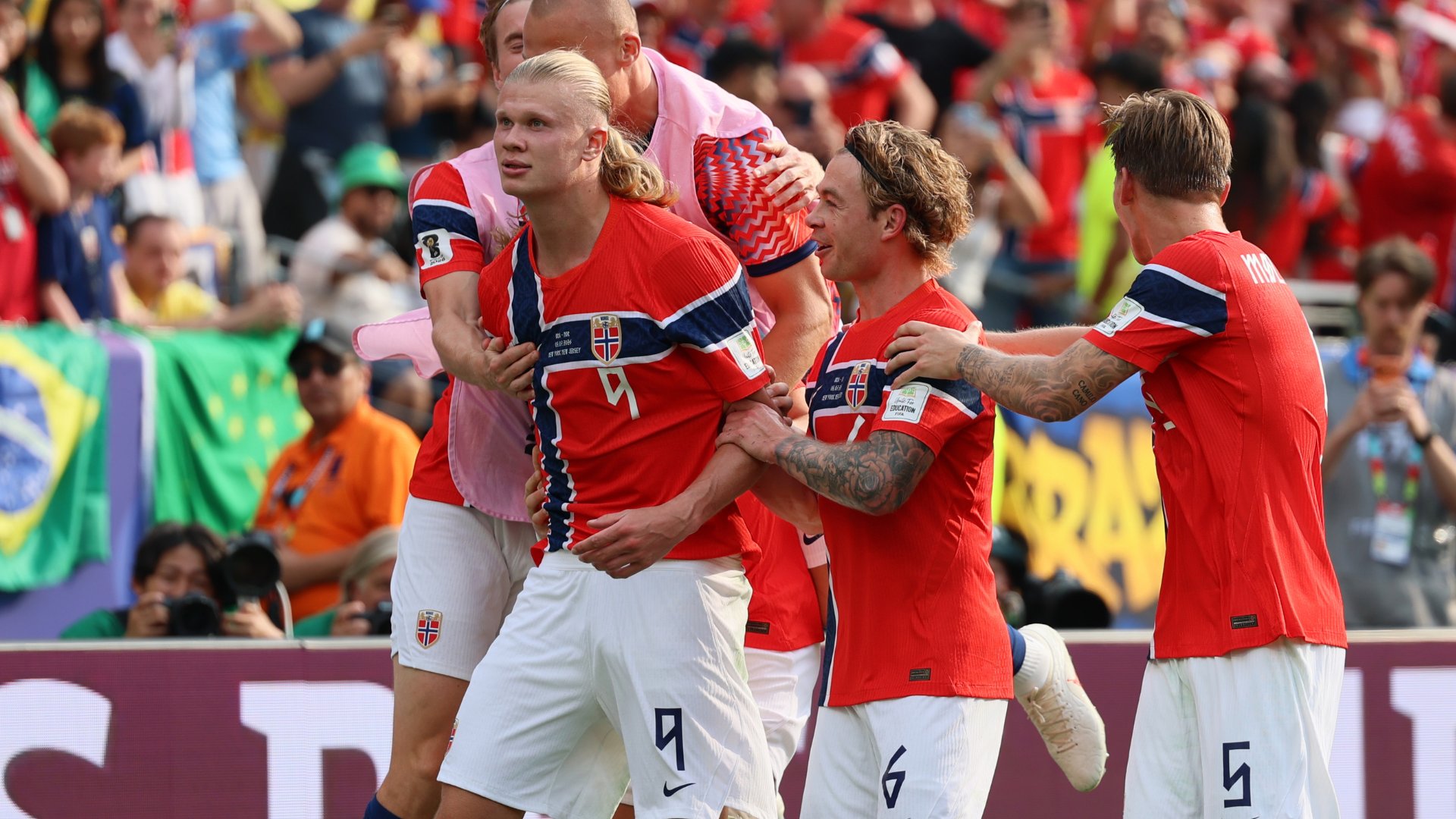 A blonde soccer player in a red jersey with number 9 celebrates with teammates in matching red kits, lifting him by the legs in front of stadium crowds.