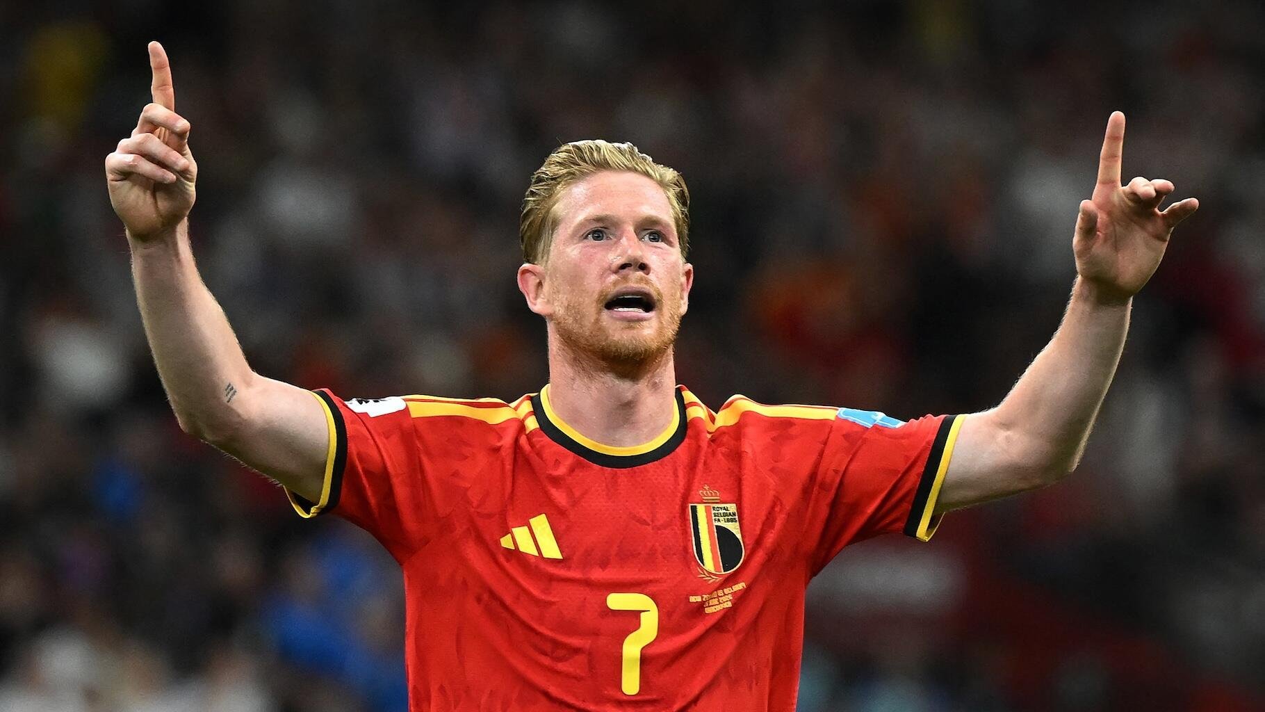 A blonde soccer player in a red Belgium jersey with the number 7 raises both arms, pointing skyward in celebration before a crowd.
