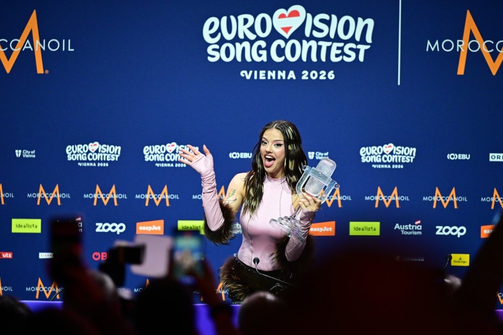 A woman in a pink top holds a glass microphone trophy and gestures excitedly at a Eurovision Song Contest Vienna 2026 press backdrop.