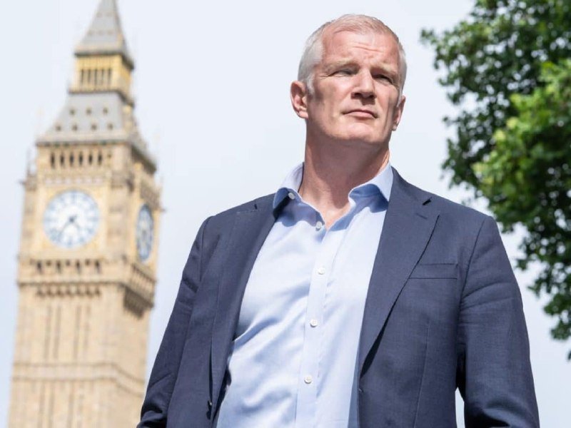 A gray-haired man in a blue blazer stands outdoors with the Big Ben clock tower and a green tree visible in the background.