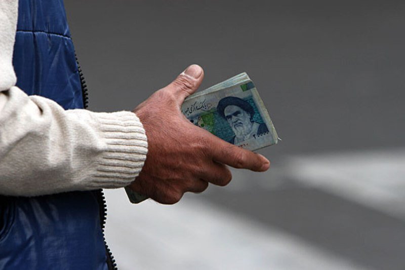 A person wearing a blue jacket and cream sweater holds folded Iranian rial banknotes in their hand against a gray background.