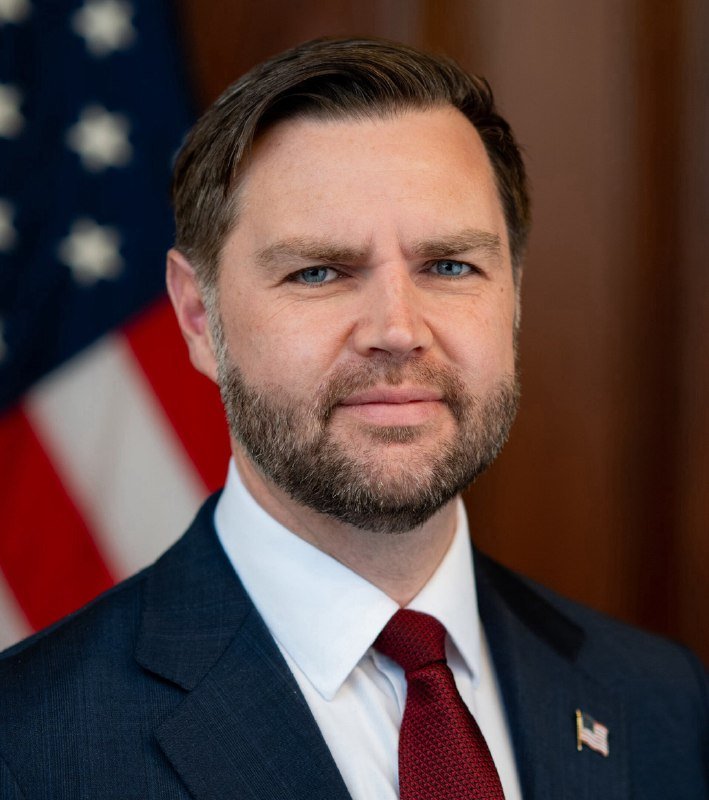 Official portrait of a bearded man in a dark suit, white shirt, and red tie, with an American flag visible in the background.