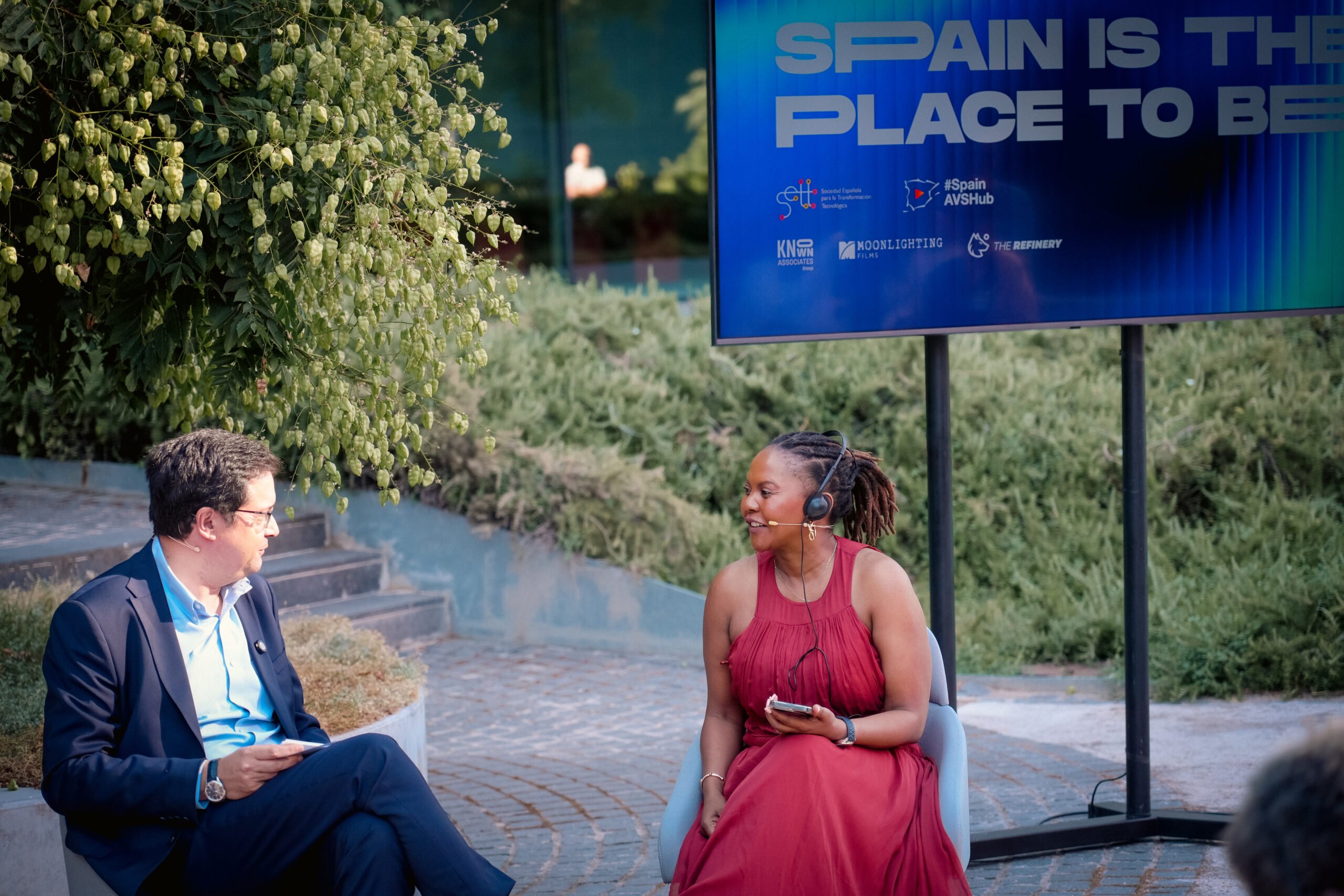 Two people sit outdoors on chairs in conversation, both wearing headset microphones, with a screen behind them reading "SPAIN IS THE PLACE TO BE."