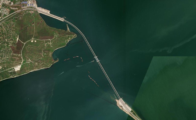 Satellite image showing a long bridge spanning across dark green water, connecting a landmass with visible agricultural plots and buildings to the opposite shore.