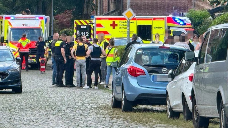 Emergency responders and police officers gather on a cobblestone street near German Red Cross ambulances and parked vehicles.