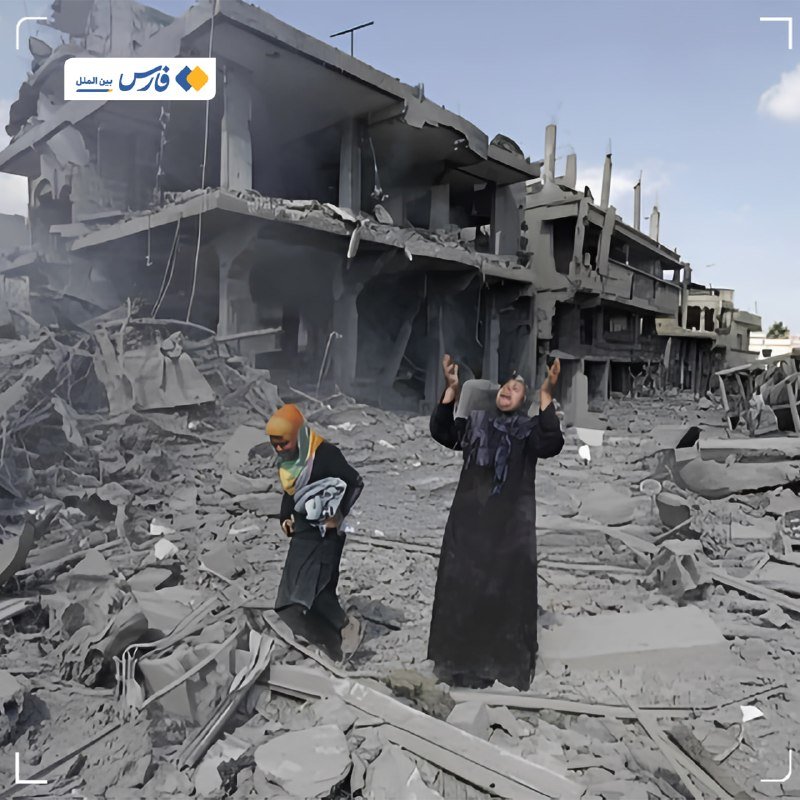 Two women stand amid extensive rubble and concrete debris in front of a heavily damaged multi-story building.