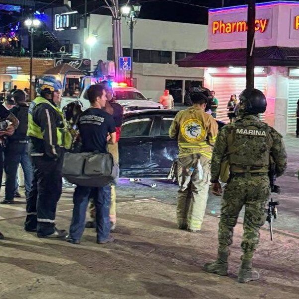 Emergency responders, including a MARINA soldier and firefighters, gather around a damaged car at night outside a Pharmacy under bright streetlights.