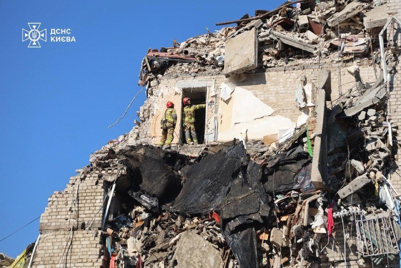 Two rescuers in tan uniforms and red helmets stand in the doorway of a heavily damaged multi-story brick building surrounded by rubble, with a blue logo and "ДСНС КИЄВА" text in the corner.