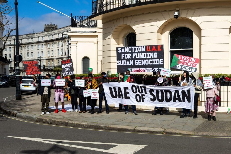 Demonstrators hold a "UAE OUT OF SUDAN" banner and signs reading "Sanction U.A.E for Funding Sudan Genocide" and "Eyes on Genocide in Sudan" outside a cream-colored building.