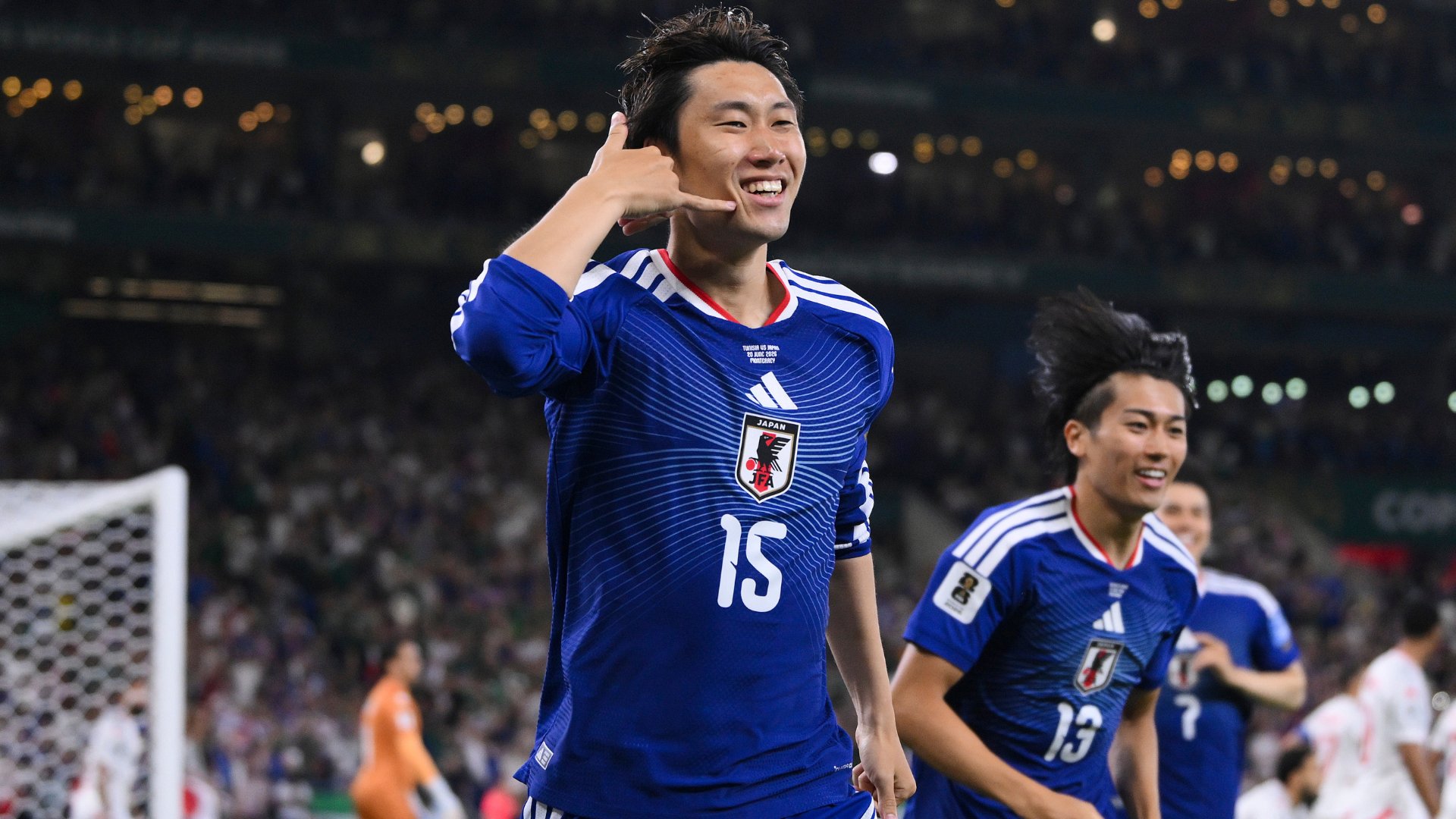 A young soccer player with bleached curly hair stands on the field wearing a red Japan national team jersey featuring the JFA crest and a FIFA World Cup 2026 qualifier patch.