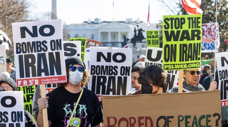 Masked and unmasked protesters hold "No Bombs on Iran" and "Stop the War on Iran" signs in front of the White House.
