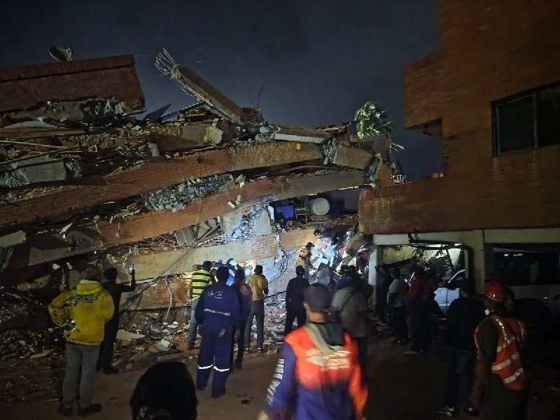 Rescue workers in red shirts search through the rubble of a collapsed concrete building, with a "TASNIM NEWS" watermark in the corner.