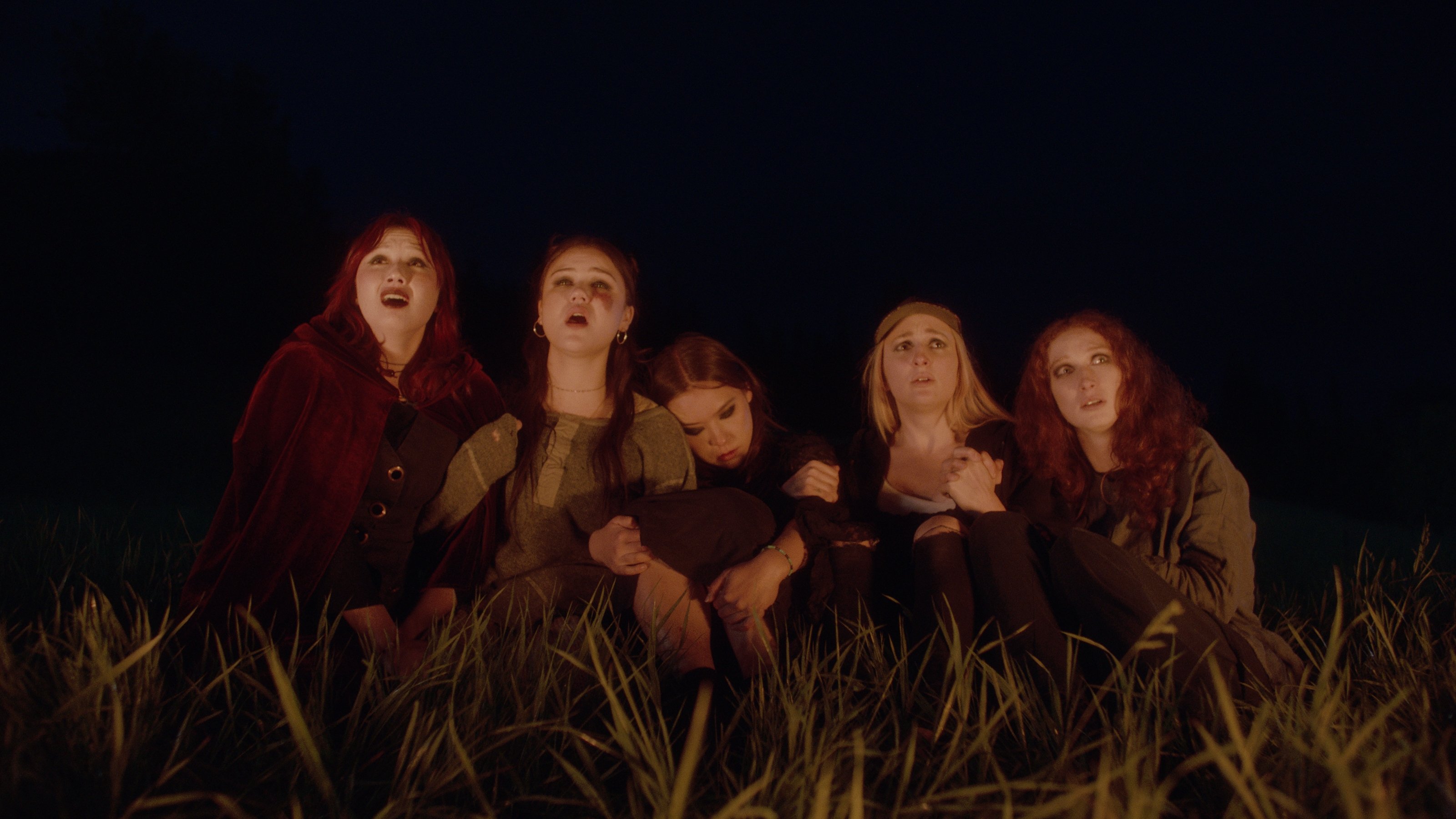 Five young women sit huddled together in tall grass at night, illuminated by a warm light, looking upward with distressed expressions.
