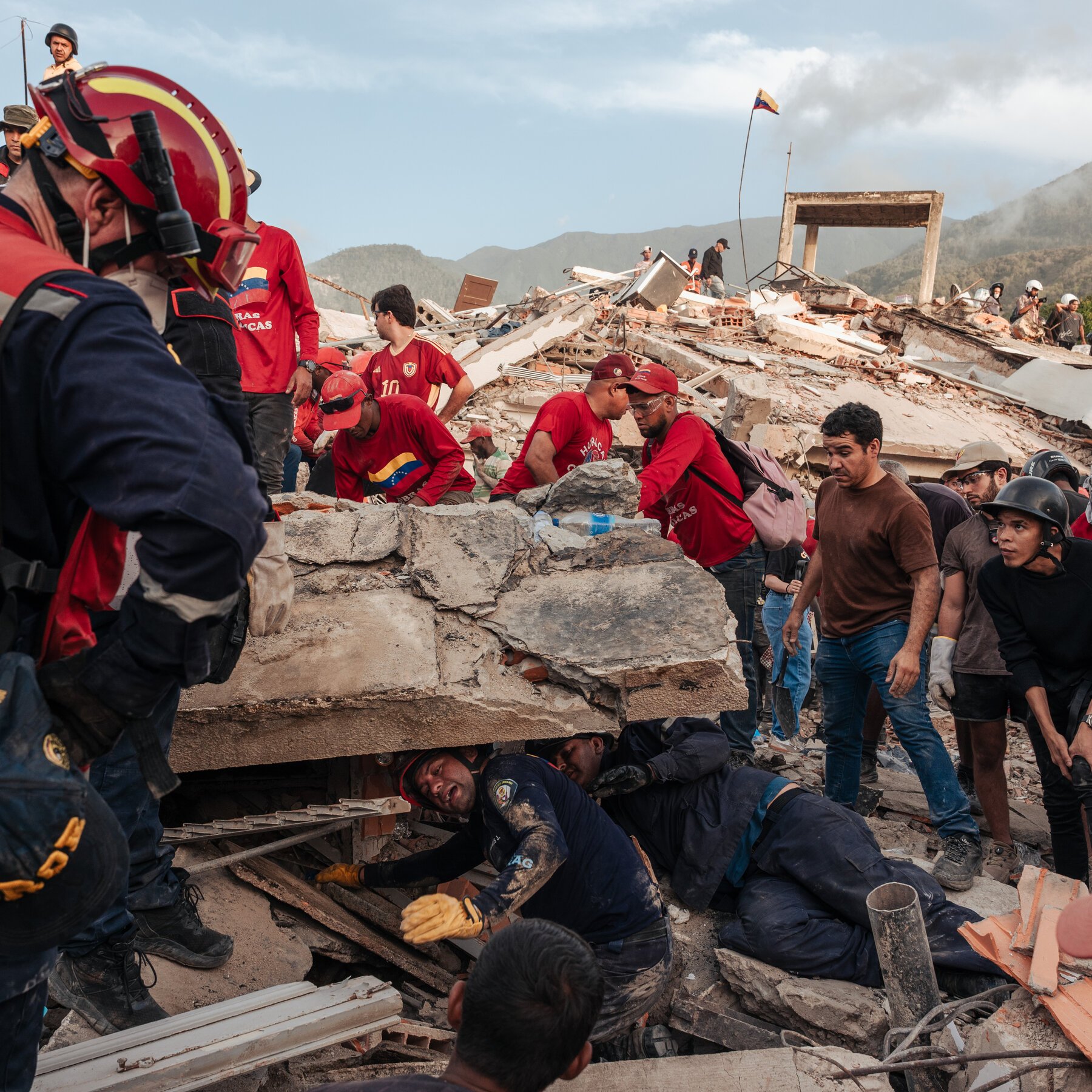 Four workers in red shirts and blue pants search through the rubble of a collapsed multi-story building at night, with a "TASNIM NEWS" watermark visible.