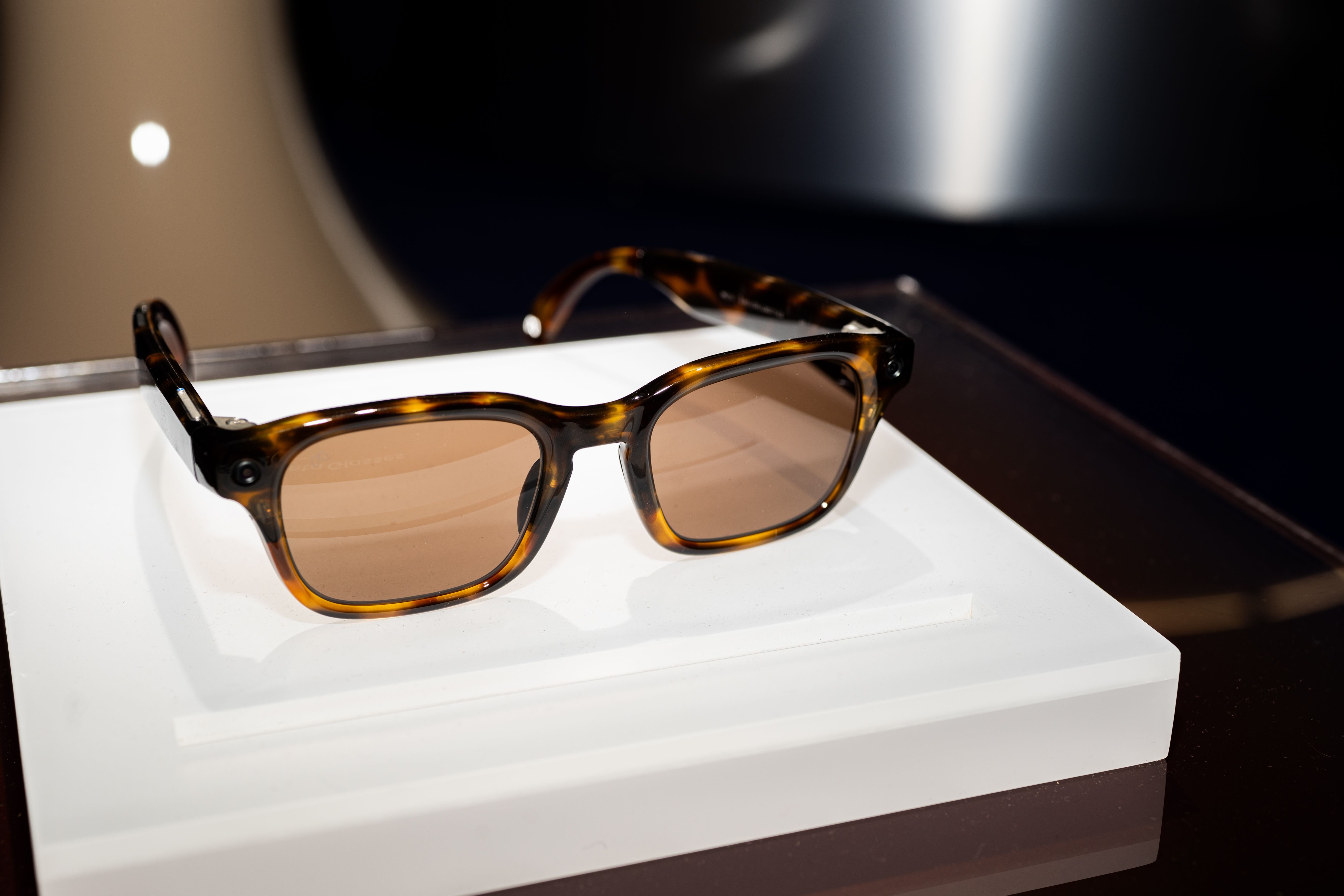 A pair of tortoiseshell smart glasses with tinted lenses rests on a white display platform in a dimly lit showcase setting.