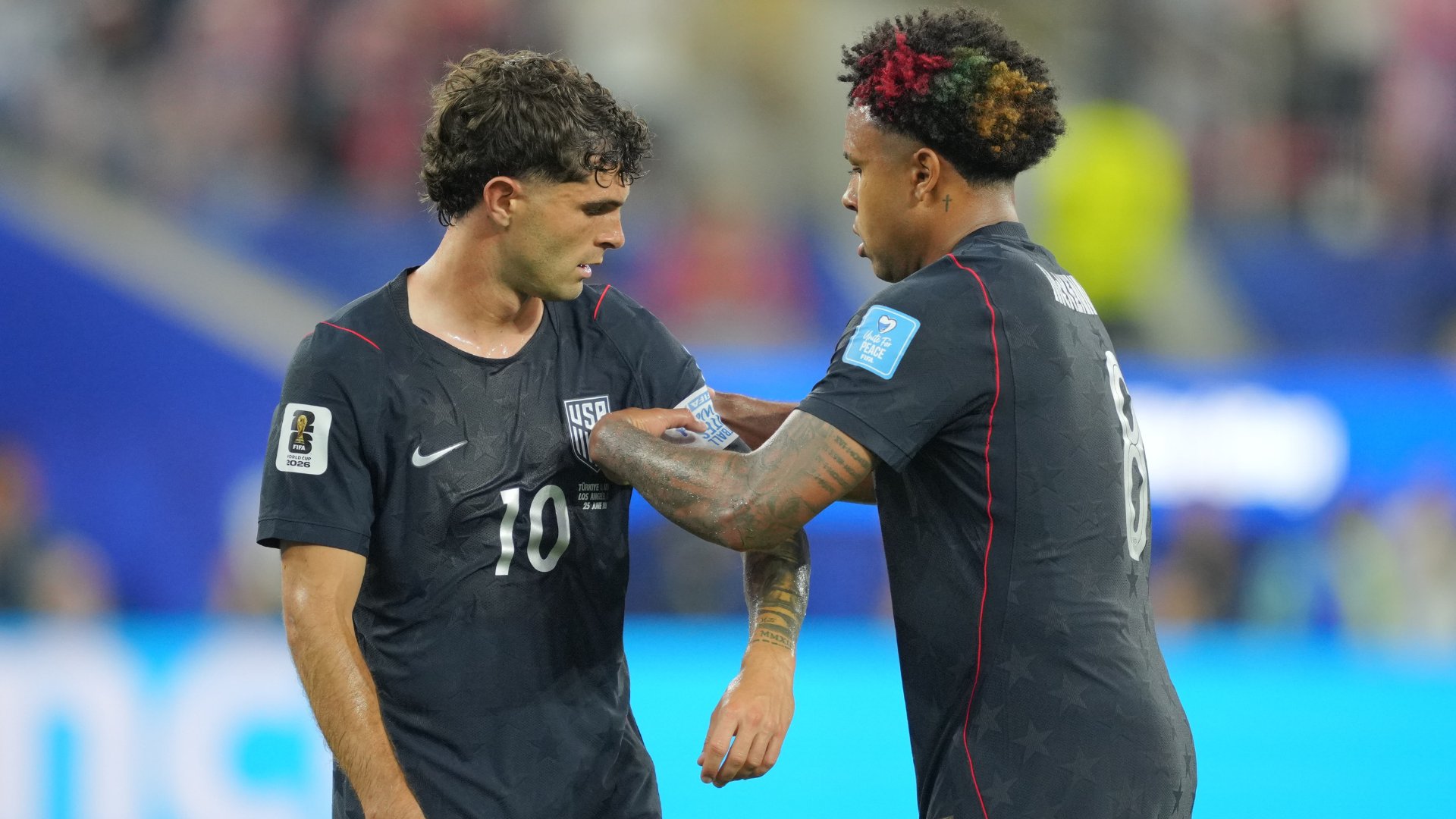 Two soccer players in dark USA national team jerseys stand face-to-face on a field, with the player on the right adjusting the captain's armband on the left player's sleeve.