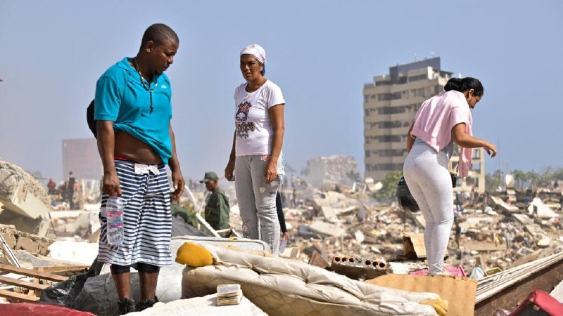 Five rescue workers in red shirts and caps sift through a large pile of rubble from a collapsed building at night.