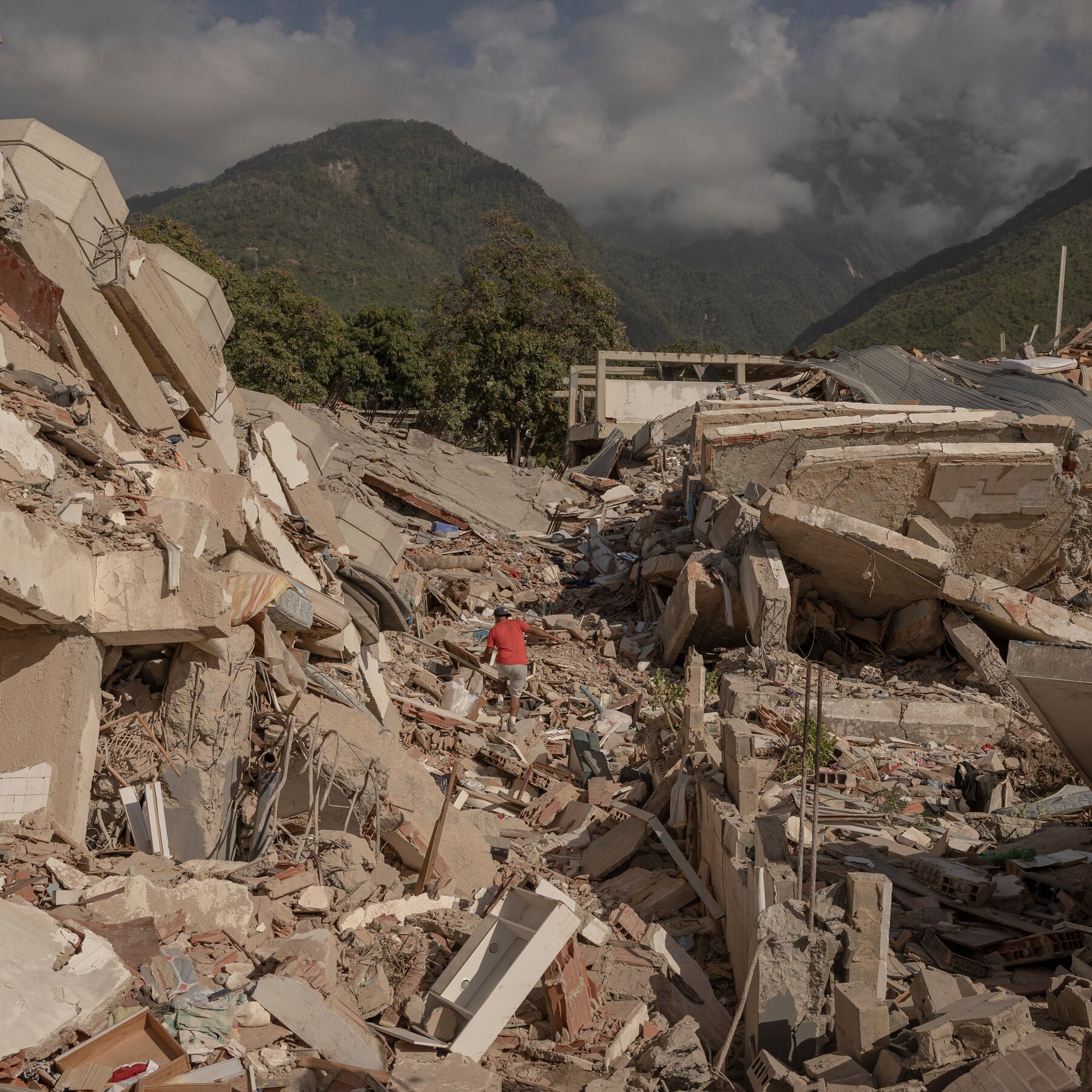 A man in a red shirt walks through massive piles of collapsed concrete and debris from destroyed buildings, set against a backdrop of cloud-shrouded mountains.