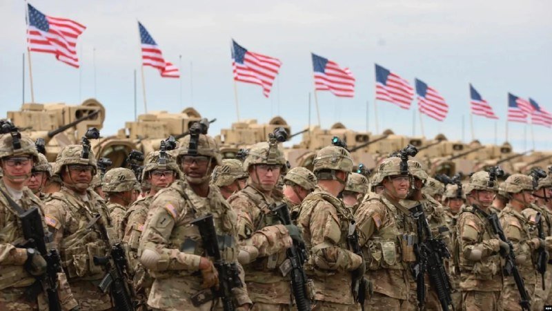 Soldiers in camouflage uniforms and helmets holding rifles stand in formation, with multiple American flags and military vehicles visible in the background.