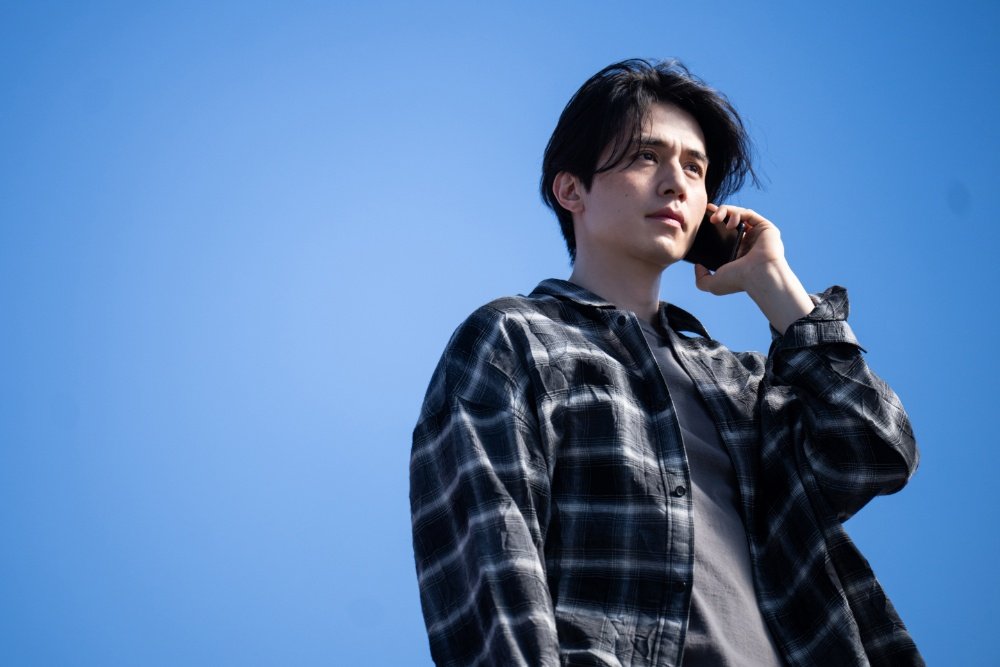 A young man with dark hair, wearing a black-and-white plaid shirt over a gray tee, holds a phone to his ear against a clear blue sky.