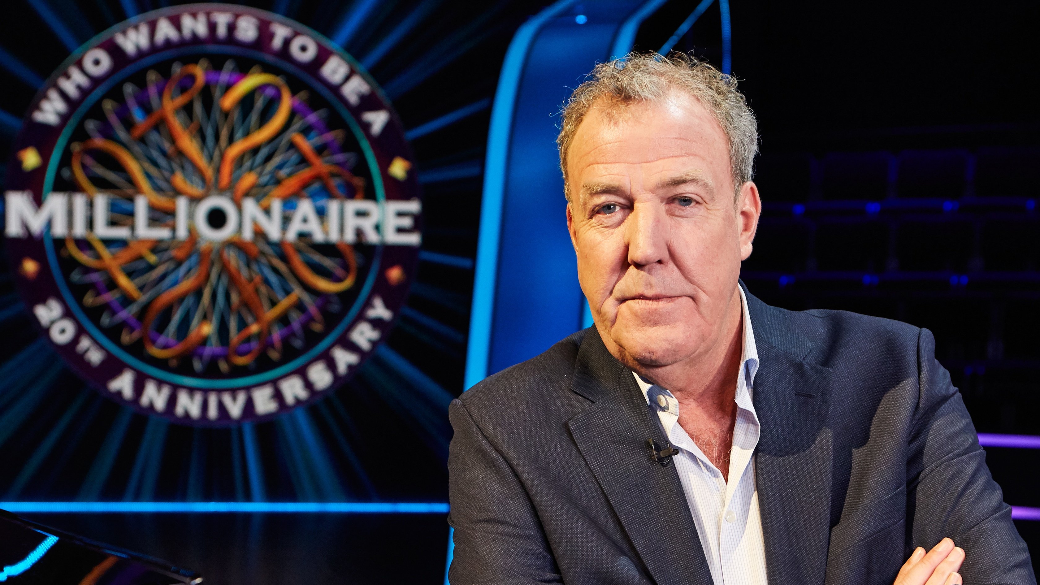A grey-haired man in a dark suit jacket and open-collared shirt sits with arms crossed beside a glowing "Who Wants to Be a Millionaire" 20th Anniversary game show logo.