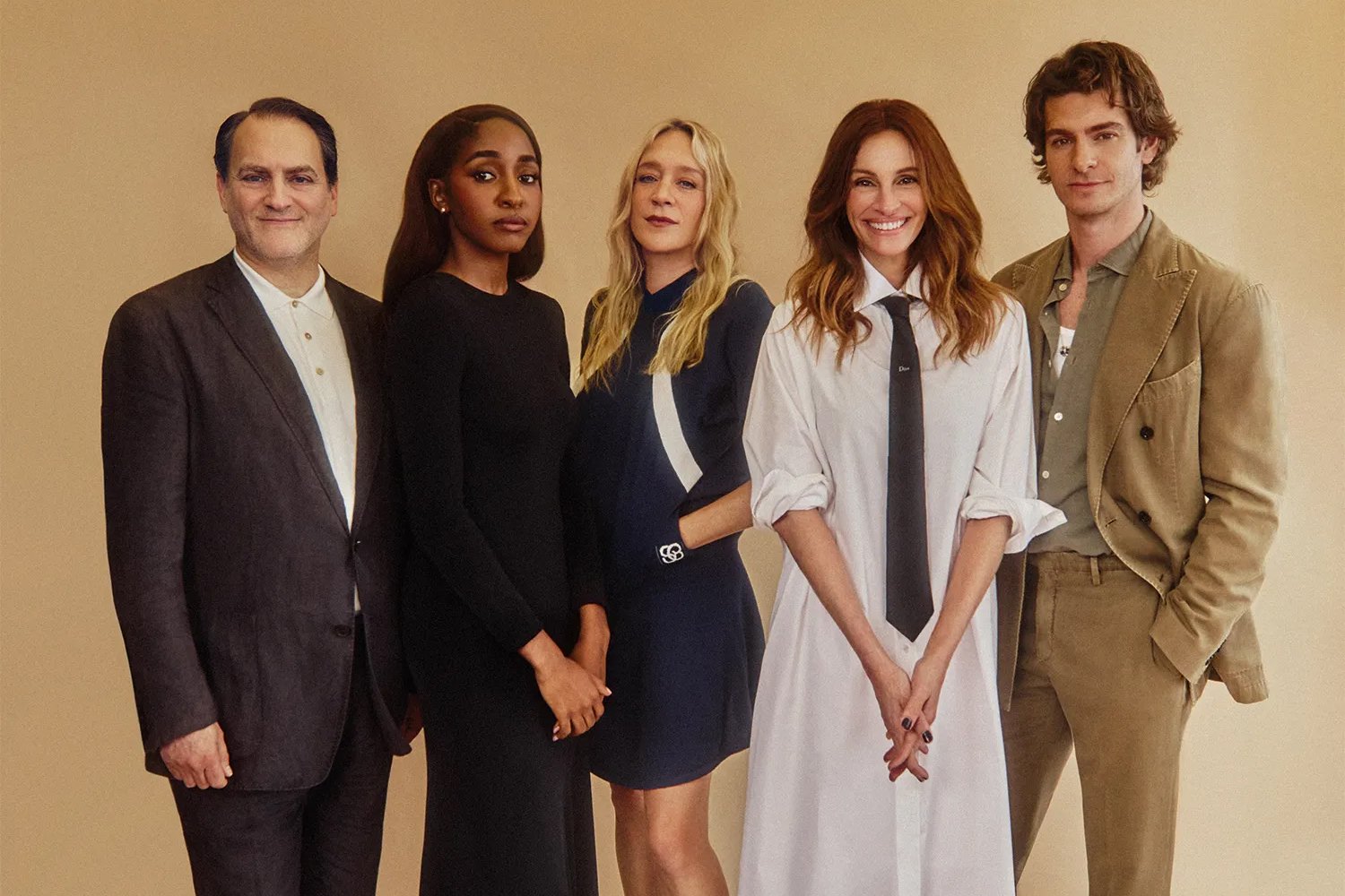 Five people pose together against a beige backdrop, dressed in neutral-toned blazers, dresses, and button-up shirts for what appears to be a promotional photo.
