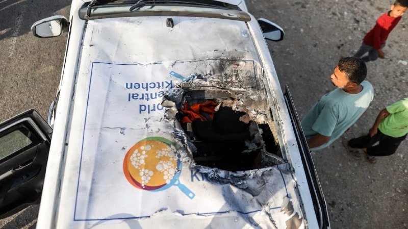A damaged white vehicle displays a World Central Kitchen logo on its burned roof, while bystanders stand nearby on a debris-strewn street.