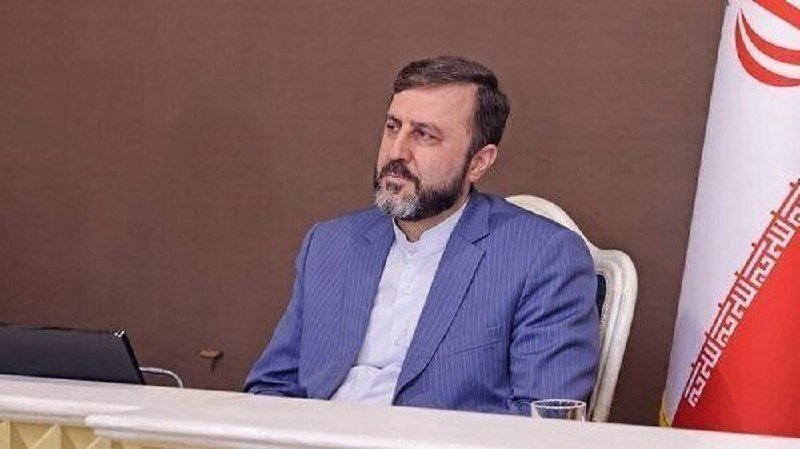 A bearded man in a blue blazer sits at a desk beside an Iranian flag, looking off to the side.