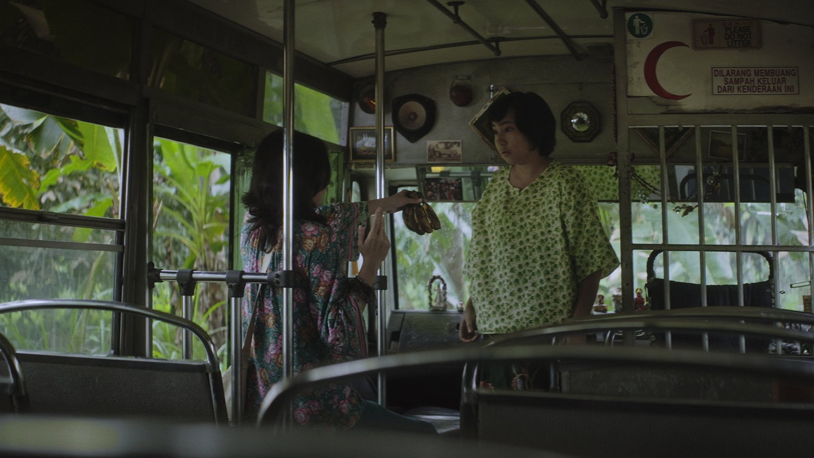 A woman in a floral dress examines an object held in her outstretched hand while another woman in a green patterned top watches, both standing inside a bus with a Red Crescent sticker visible.