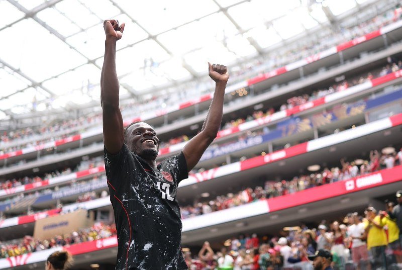 A smiling soccer player wearing a black jersey with the number 12 raises both arms in celebration inside a crowded stadium.