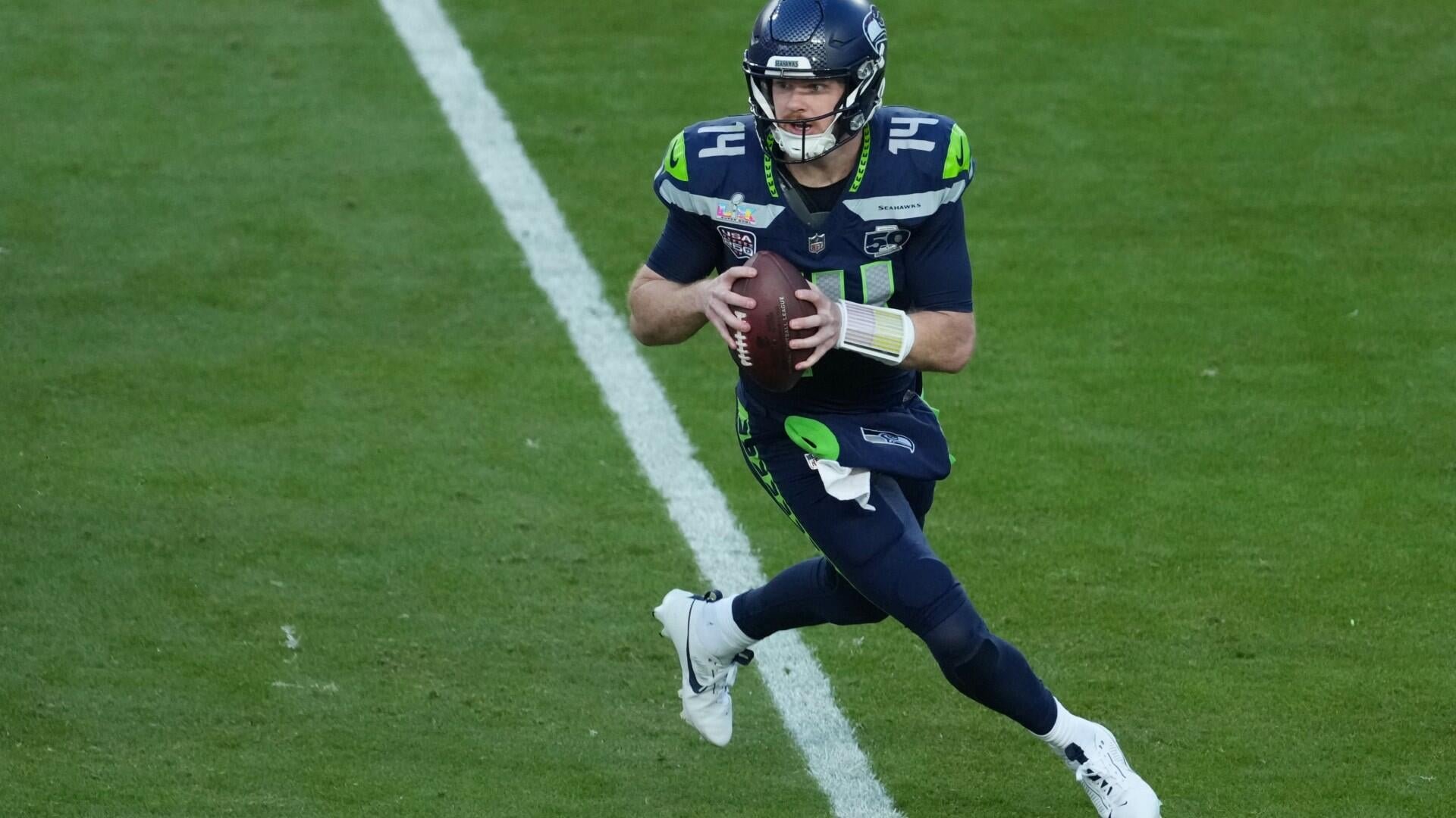 A football player in a navy blue and neon green Seahawks uniform runs across a grass field, gripping a football in both hands.