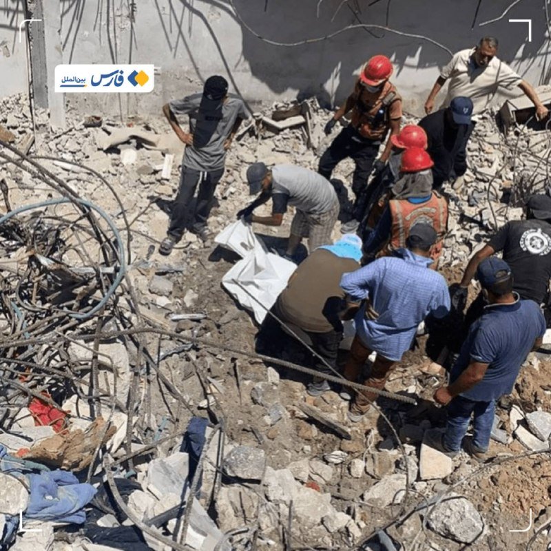 Rescue workers in helmets and vests gather around a white body bag amid concrete rubble and exposed rebar at a building collapse site.