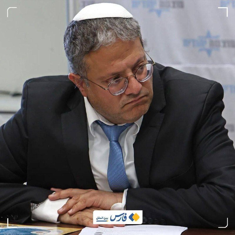A gray-haired man wearing glasses, a kippah, dark suit, and blue tie sits at a table looking down at papers.