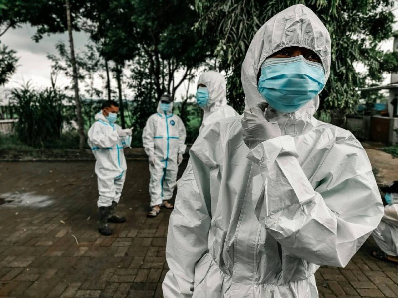 Three workers in yellow protective suits, white aprons, and face shields pour contents from a bag into buckets near a weathered building's steps, behind orange safety fencing.