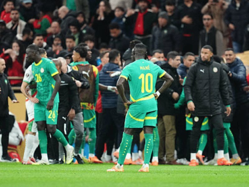 A Senegalese football player wearing the number 10 jersey with "MANÉ" stands with hands on hips on the sideline, surrounded by teammates, staff, and a crowd of spectators.