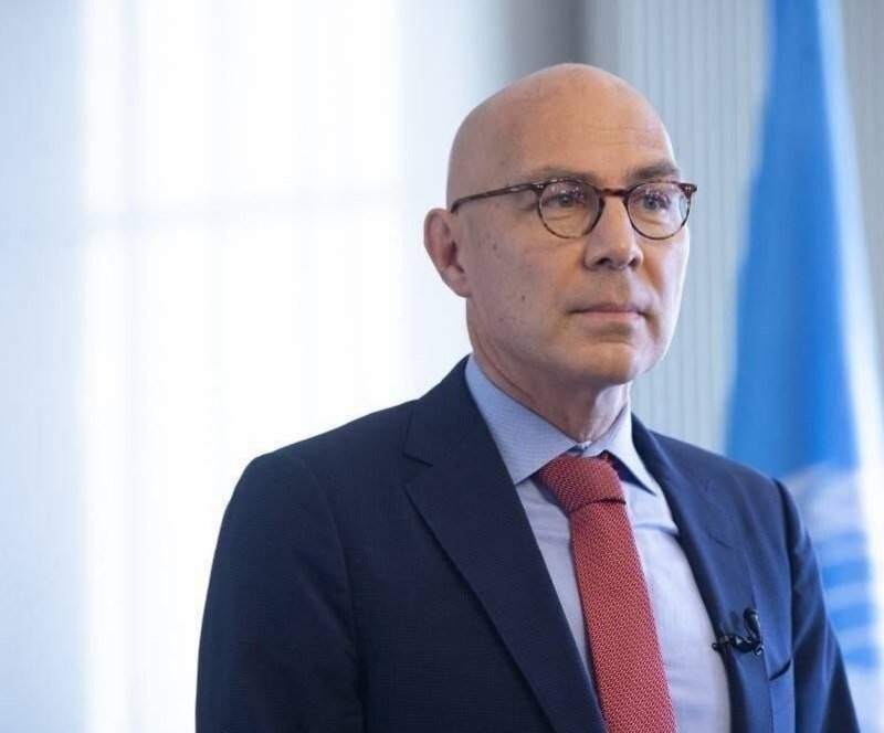 A bald man wearing glasses, a navy suit, and red tie speaks in front of a blue and white flag and light curtains.