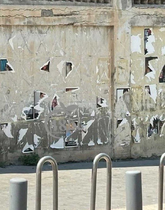 Torn and peeling posters cover a weathered concrete wall, with metal bike racks and bollards visible in the foreground on a paved surface.