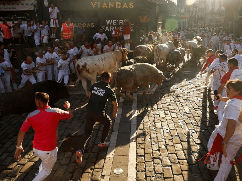 Crowds of people in white clothing with red sashes run alongside a herd of bulls down a cobblestone street, with some wearing "PASTORES" shirts.
(28 words)