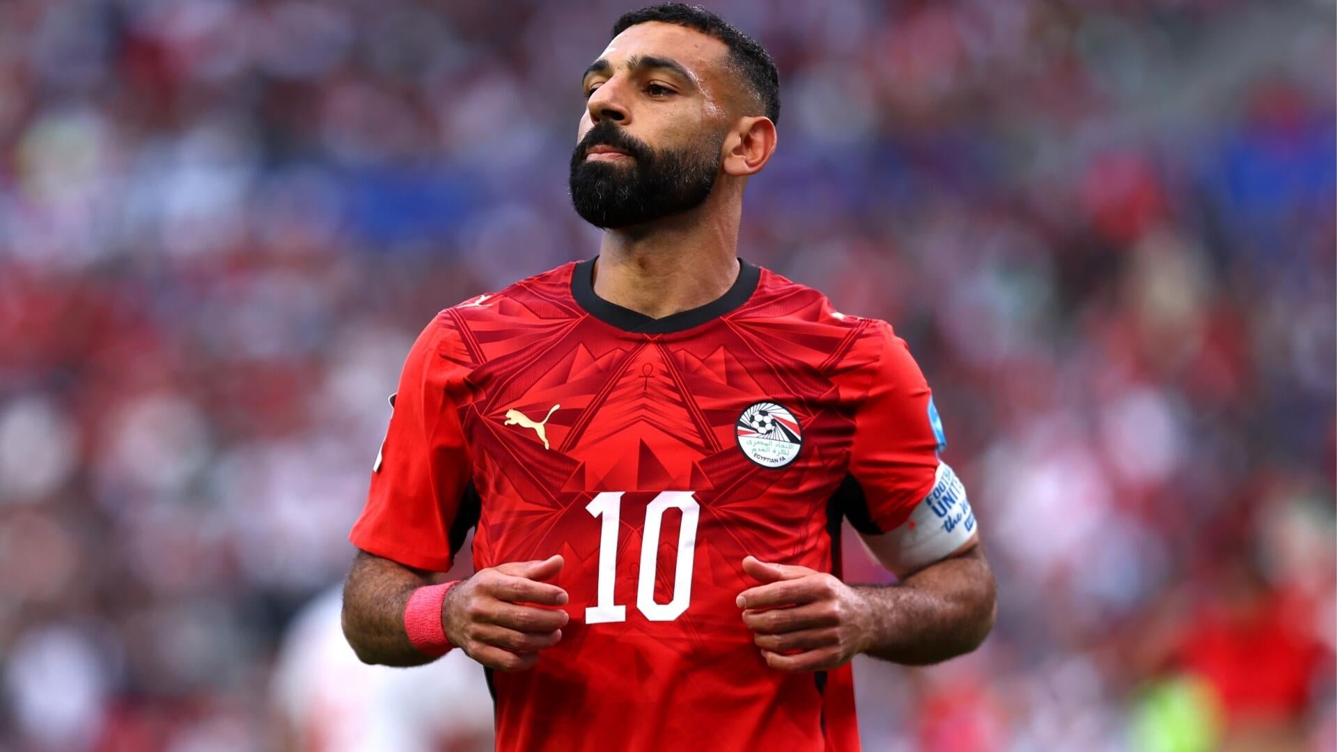 A bearded soccer player in a red jersey with the number 10 and an Egyptian Football Association crest stands in a stadium, wearing a captain's armband.