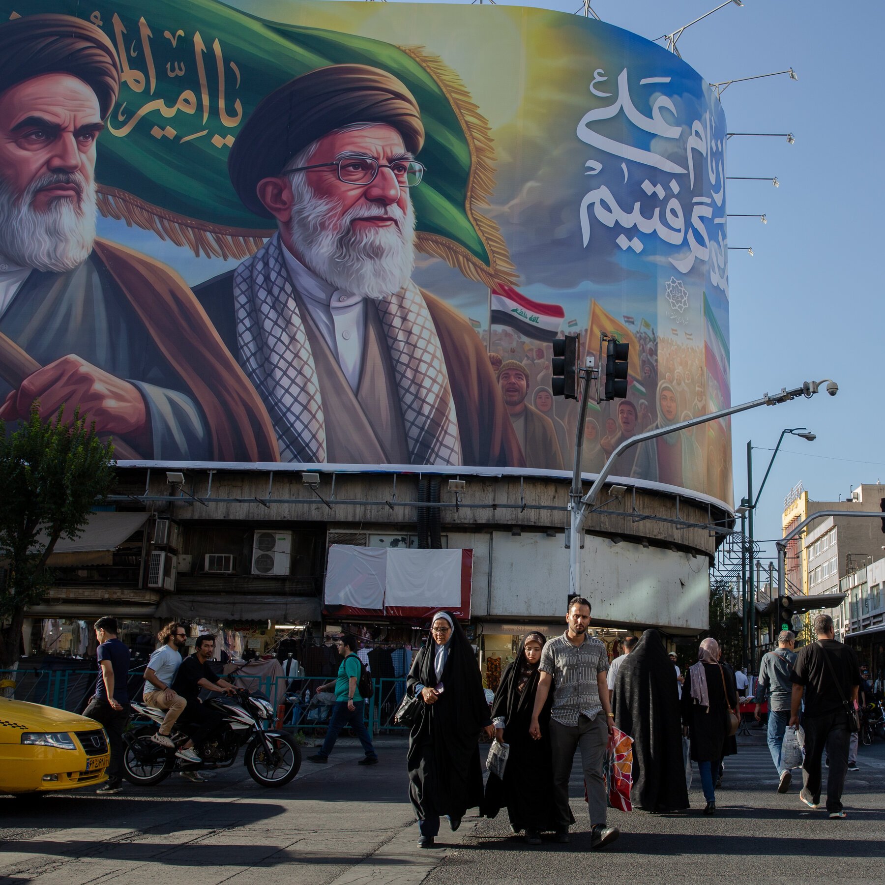A man in glasses and a dark suit stands at a podium bearing an Iranian emblem, with an Iranian flag and a blue map backdrop reading "...Y OF ...FFAIRS."