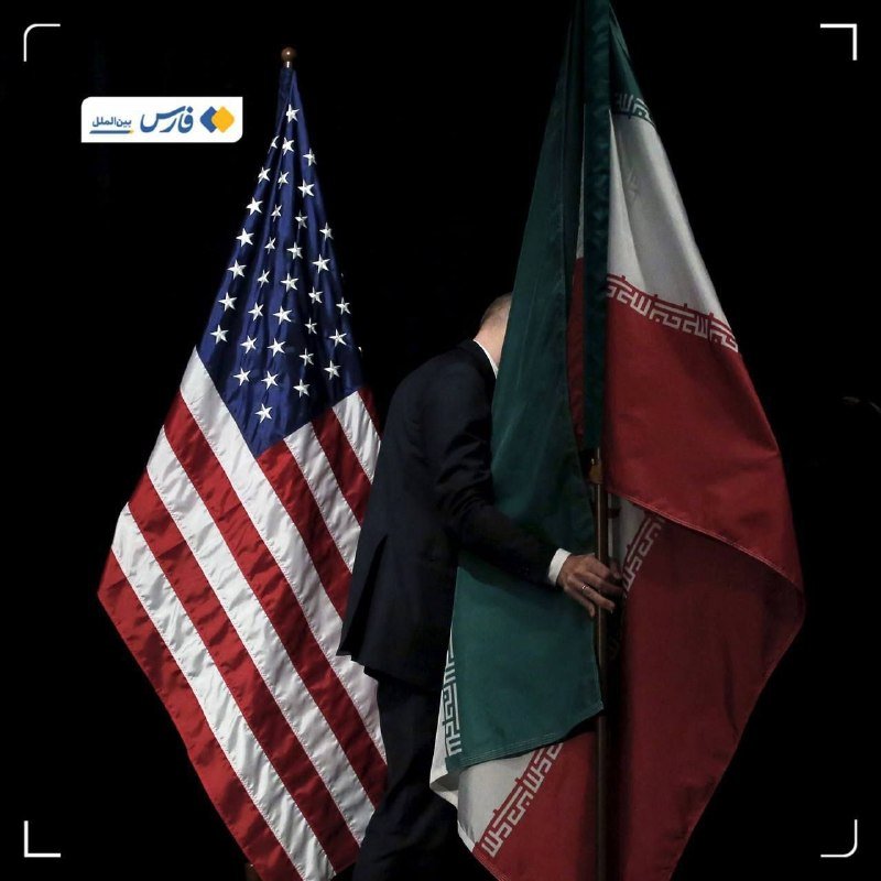 A man in a dark suit stands between the flags of the United States and Iran against a black background, his head turned away.