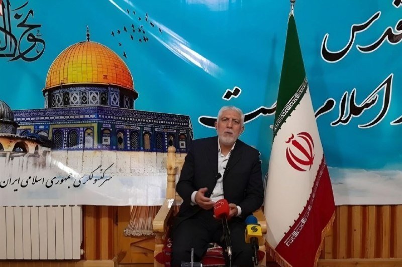 A bearded man in a dark suit sits before microphones, speaking in front of an Iranian flag and a backdrop depicting the Dome of the Rock with Persian script.