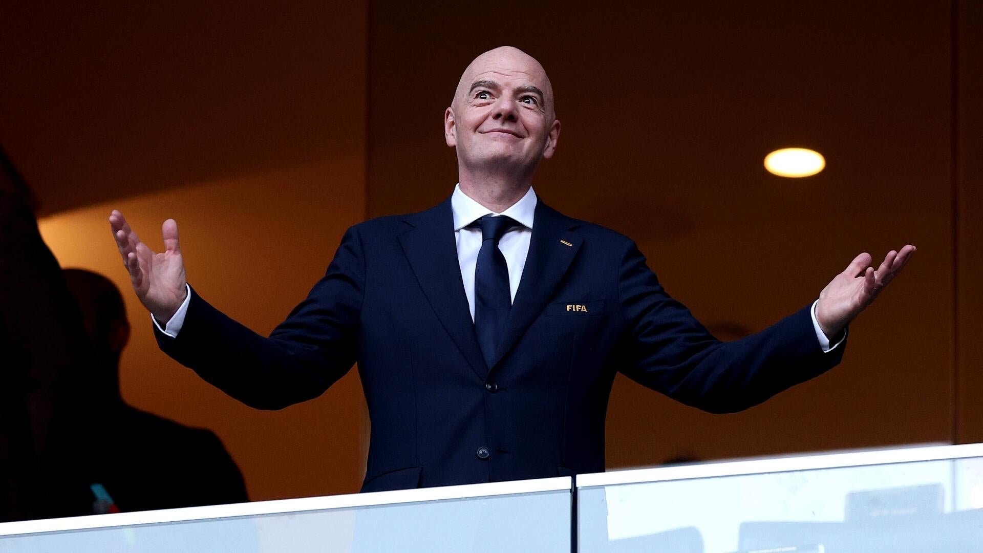 A bald man in a dark suit with a "FIFA" name badge raises both arms with open palms, looking upward with a smile from behind a glass barrier.