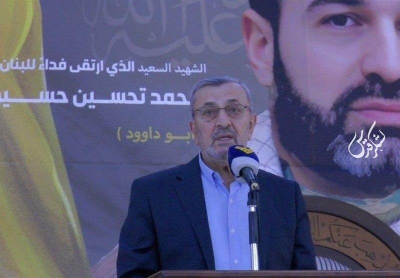 A bespectacled man in a dark suit speaks into a microphone at a podium, standing before a large banner displaying Arabic text and a portrait of a bearded man.