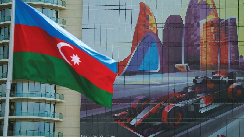 The Azerbaijani flag waves in the foreground of a building displaying a large mural of a Formula One race car and futuristic architecture.