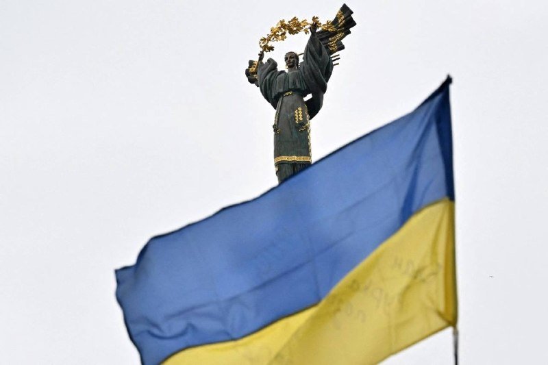 A blue-and-yellow flag waves in the foreground with a gold-accented statue of a robed figure holding a wreath, set against a pale gray sky.
